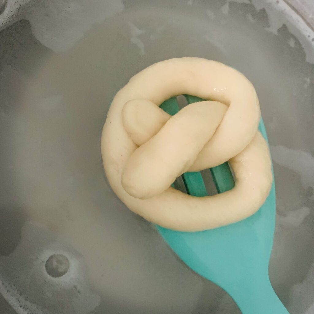 Bird's eye view: unbaked soft pretzel dough on an aqua spatula being removed from a baking soda bath.