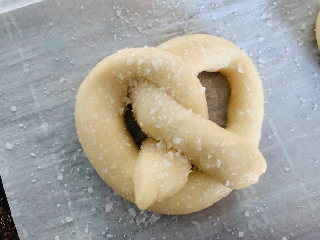 Bird's eye view: unbaked, salted soft pretzel dough on a baking sheet with white parchment paper.