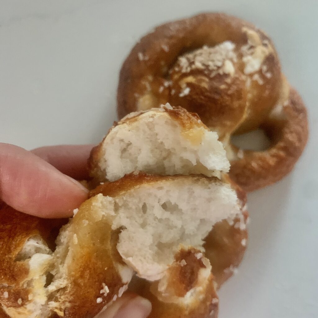 Hand showing the fluffy, soft inside of a broken open soft pretzel, with a whole pretzel in the background.