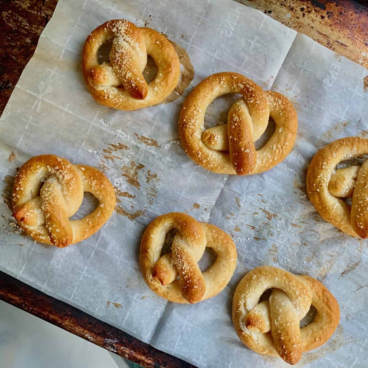 Birds Eye view: 6 baked gluten-free soft pretzels with salt on a baking sheet with parchment paper.