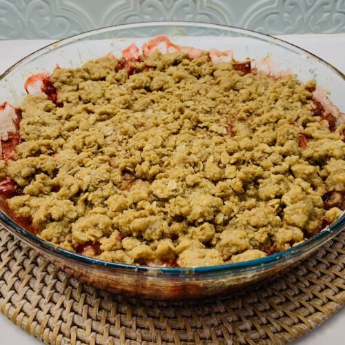 Gluten-free strawberry rhubarb crisp in a glass pie dish on a round, straw-colored charger.