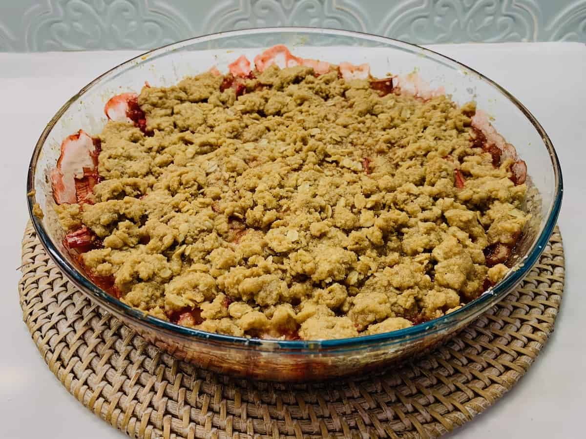 Gluten-free strawberry rhubarb crisp in a glass pie dish on a round, straw-colored charger.