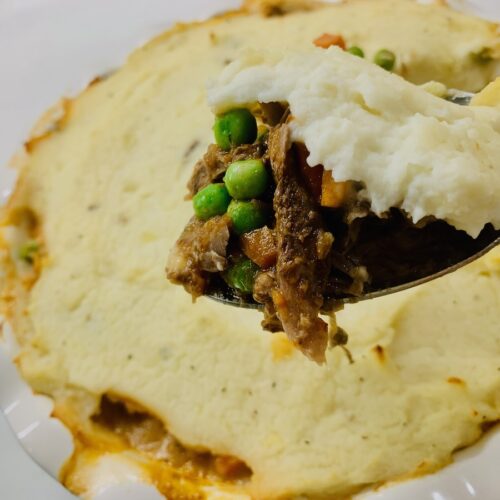 Spoonful of lamb, peas & carrots, and mashed potatoes held up with a lamb shepherd's pie in the background.