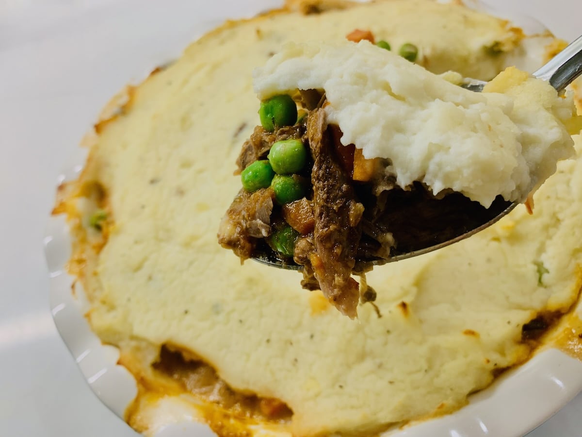 Spoonful of lamb, peas & carrots, and mashed potatoes held up with a lamb shepherd's pie in the background.