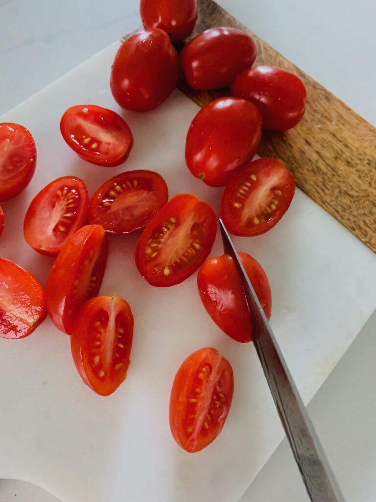 Knife slicing plum tomatoes in half on a marble/wood cutting board.
