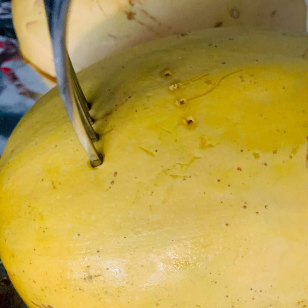 A fork piercing the skin of a yellow spaghetti squash half, with the other half in the background.