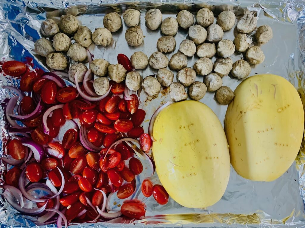 Birds Eye view: tomatoes, onions, gluten-free meatballs and 2 upside-down spaghetti squash halves on a foil-covered sheet pan.