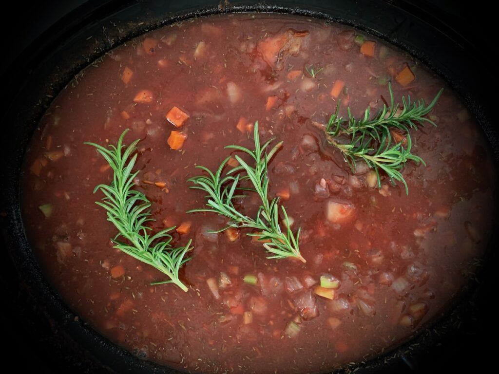 Birds Eye view: slow cooker with uncooked red sauce, visible diced vegetables, deep red sauce, topped with 3 bright green sprigs of rosemary.