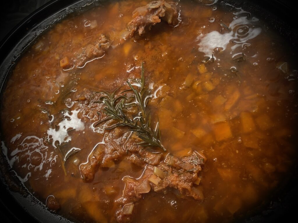 Birds Eye view: slow cooker with cooked red sauce, visible parts of lamb shanks, diced carrots, and cooked sprigs of rosemary.