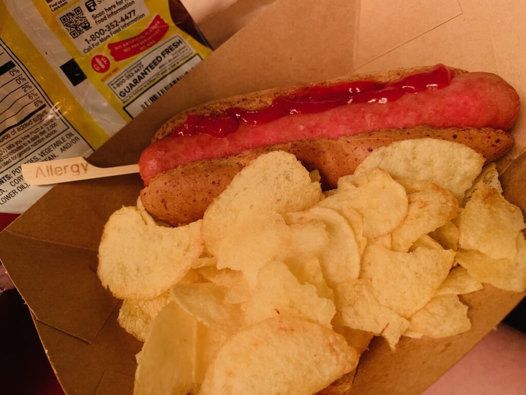 Gluten-free hot dog in a bun with ketchup and an allergy pick and potato chips.