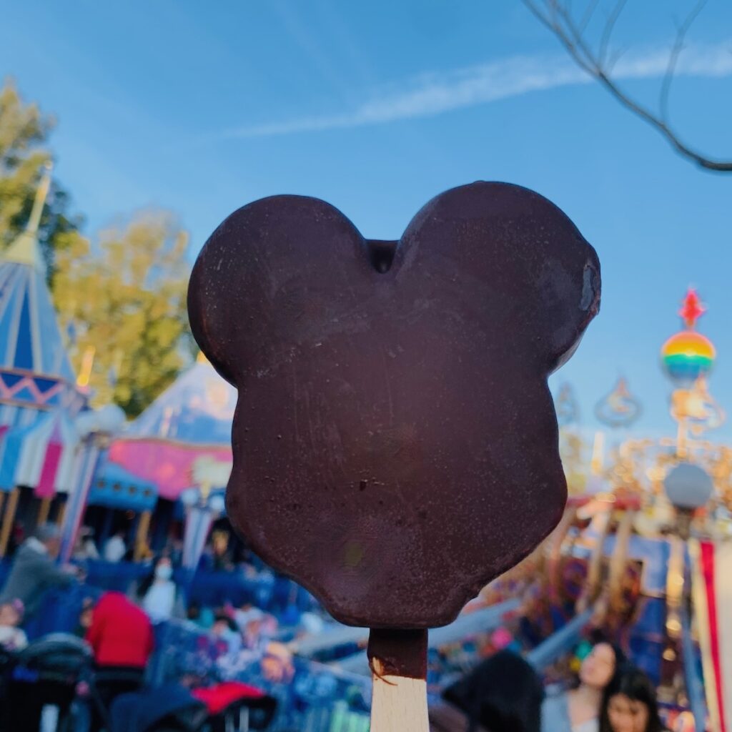 Chocolate Mickey ice cream bar held up with colorful Disney rides in the background.