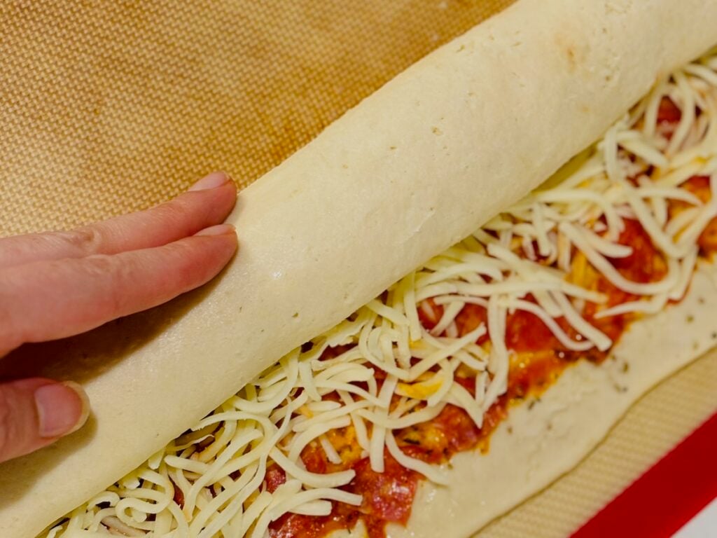 a hand rolling up a log of pizza dough, half way rolled with visible sauce, pepperoni pieces and shredded white cheese and a strip of dough without toppings along the edge