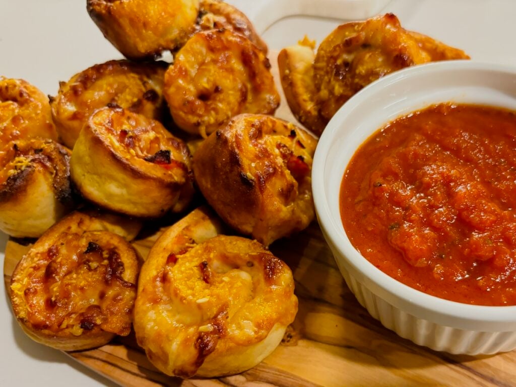 A pile of gluten-free pizza rolls and partly visible white round ramekin of tomato sauce on a wooden cutting board