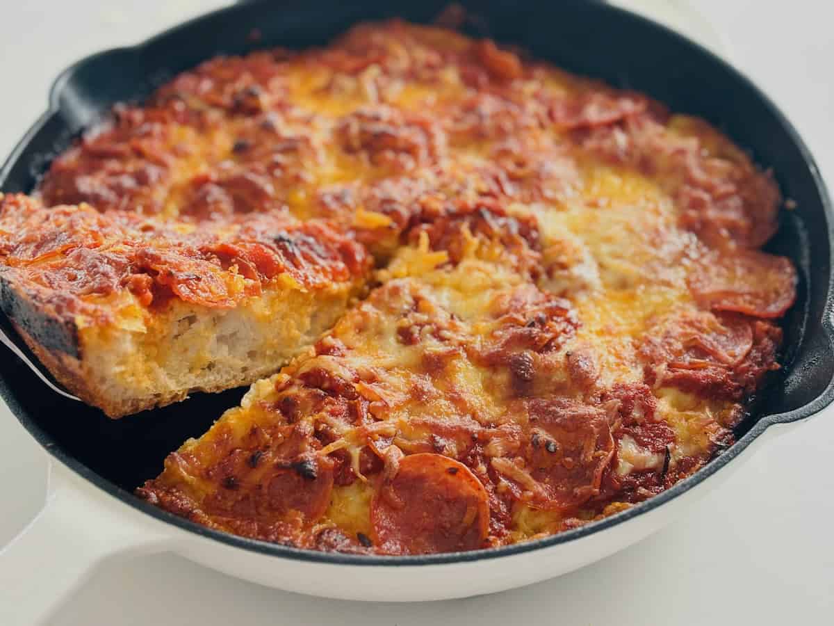 A slice of thick crust pan pizza being lifted out of a cast iron skillet with a metal cake lifter.