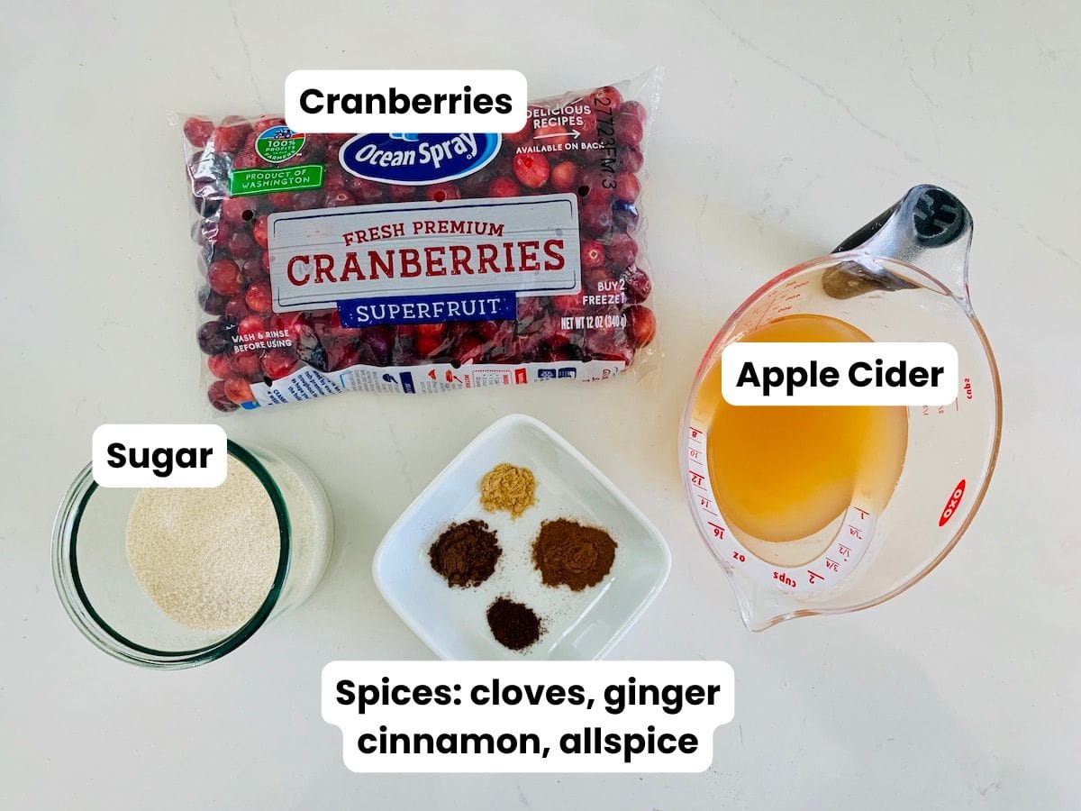 Ingredients on a white counter: cranberries, apple cider, spices (cloves, ginger, cinnamon, allspice), and sugar.