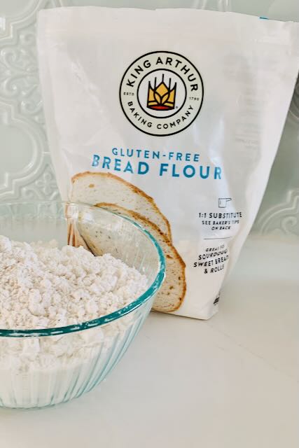A glass bowl of flour in front of a bag of King Arthur Gluten-Free Bread Flour.
